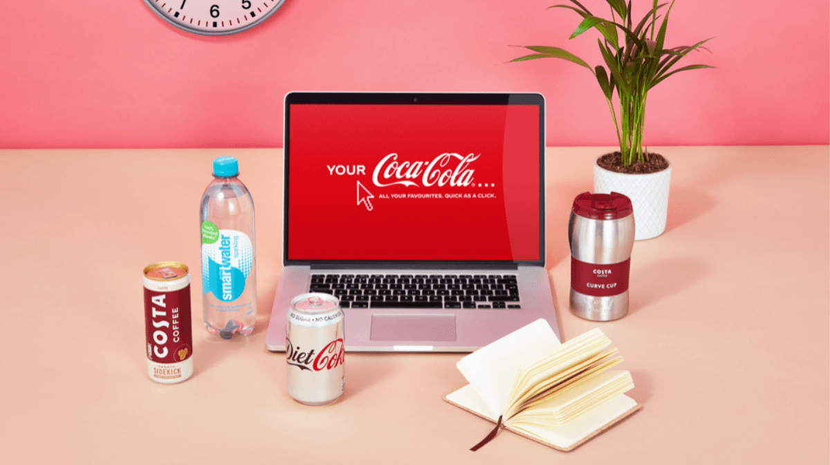 A laptop displaying the Your Coca-Cola logo, surrounded by beverages included as part of the Home-Working bundle, such as Diet Coke, Smartwater Sparkling and Costa Coffee