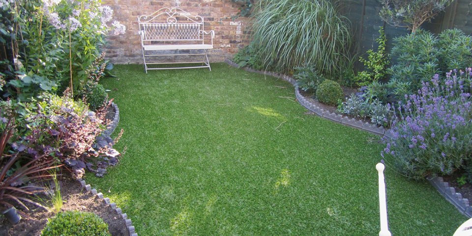 Artificial turf laid down in a garden, surrounded by a flower bed, with a white bench positioned on it