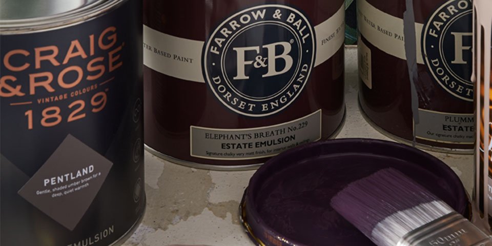 A close up image of some paint cans - varying in colour and brand - with one of the tins open and a brush resting on top of it