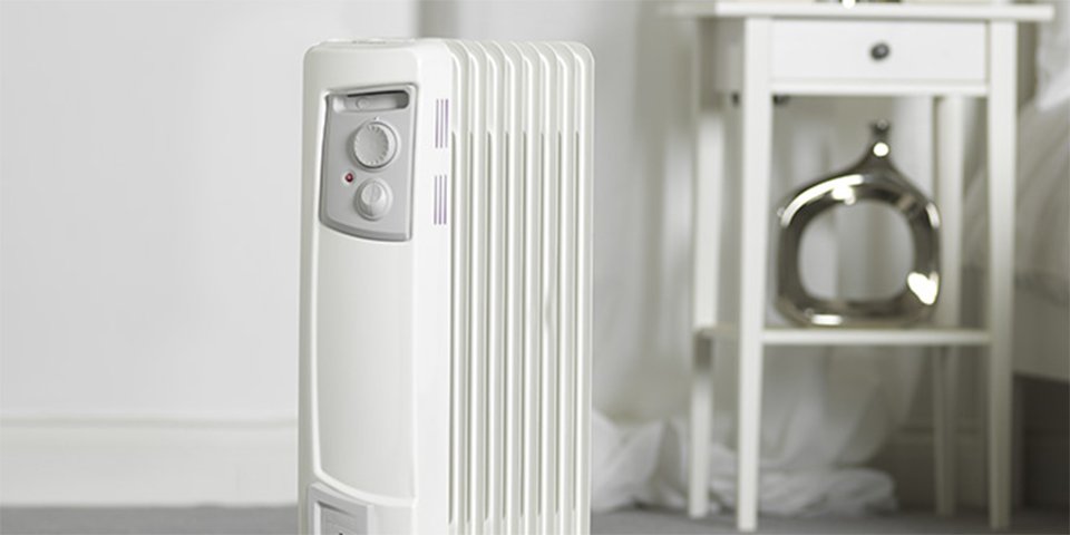 A white heater with grey dials, placed in the middle of a room near a bedside unit.