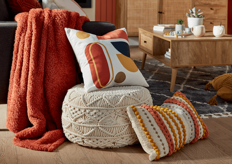 an image of orange, yellow and white patterned cushions on a woven textured footstall with an orange fluffy throw