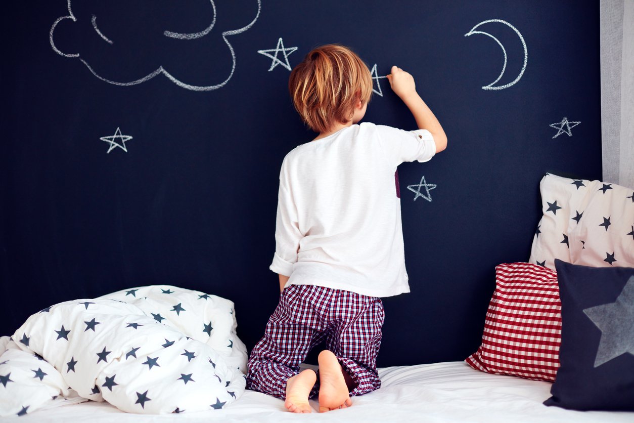An image of a child on their bed, using chalk to draw shapes on their bedroom walls