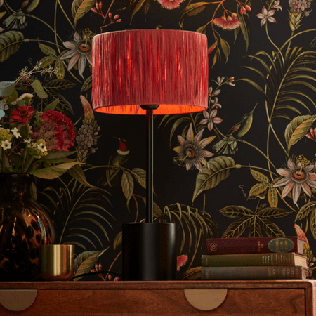 Red, ribbed lamp shade with black lamp base and patterned wallpaper in the background.