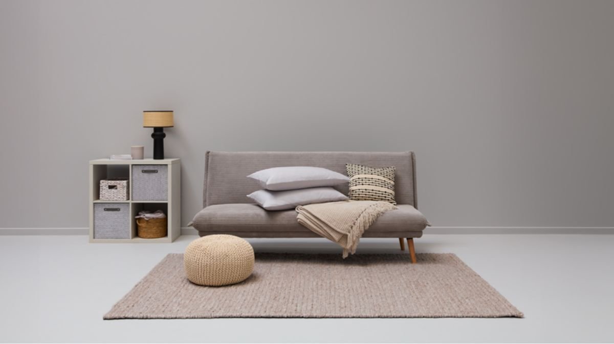 grey sofa bed decorated with soft furnishings behind a rattan rug and footstool