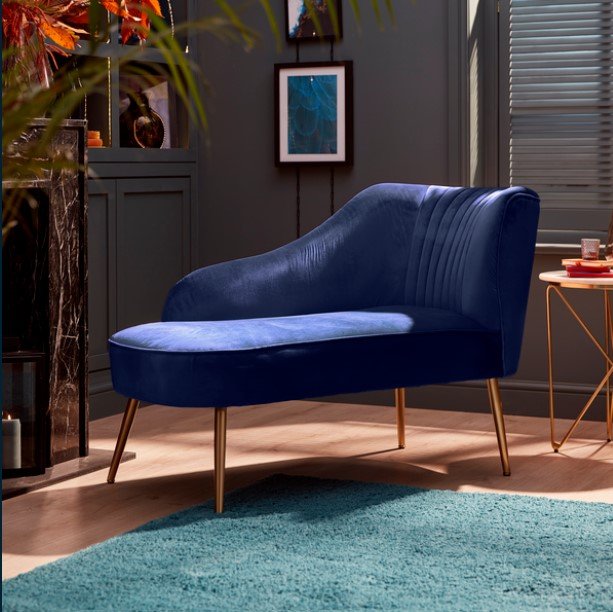 Image of a royal blue velvet modern chaise longue with brass legs positioned in a modern blue living room furnished with copper accents and a teal rug.