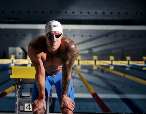 Swimmer Caeleb Dressel stood next to a pool wearing Speedos, a swim cap and goggles.