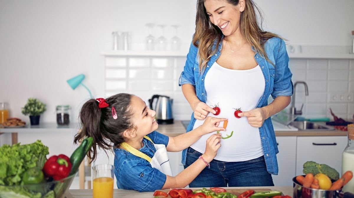 Una mamma incinta e sua figlia ridono allegramente con del cibo in mano in cucina.