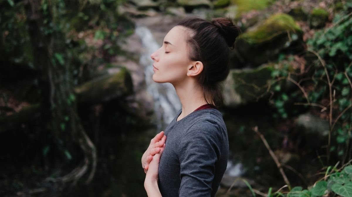 Una giovane donna in piedi respira pacificamente ed e' circondata dalla natura e da una piccola cascata 