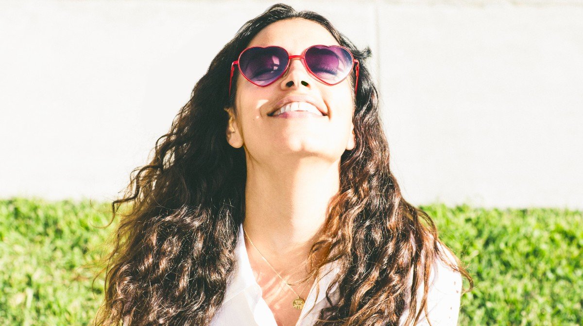 curly haired woman smiles in sunlight with heart sunglasses