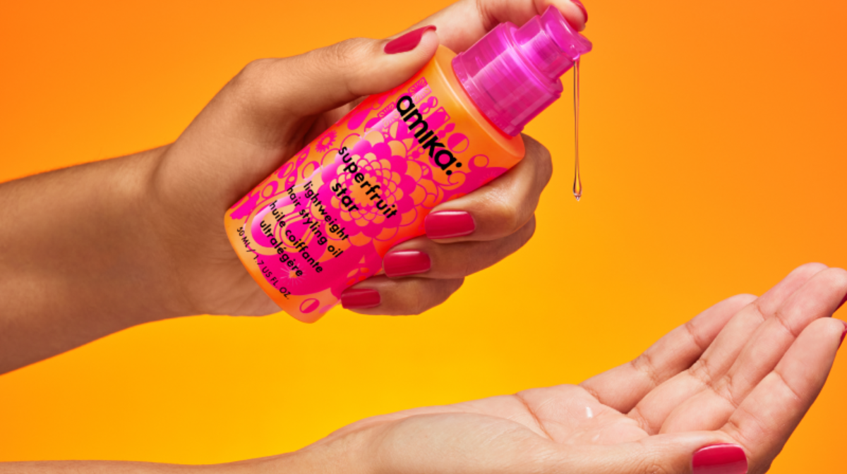 Amika Superfruit Hair Oil being dispensed from a pump into an open palm against a vibrant orange background