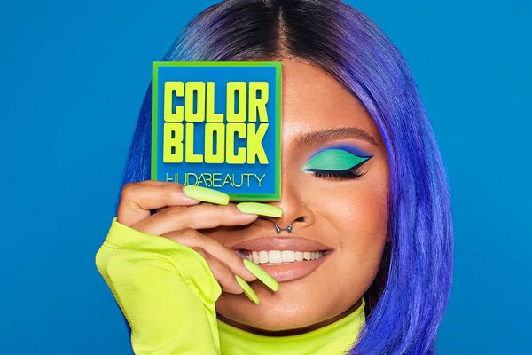 model with blue hair holding a huda beauty color block palette to her eye while the other is closed wearing green bright eyeshadow
