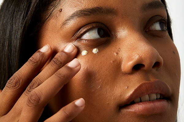 A close up image of a female model layering her skincare and applying her eye cream, in a studio setting.