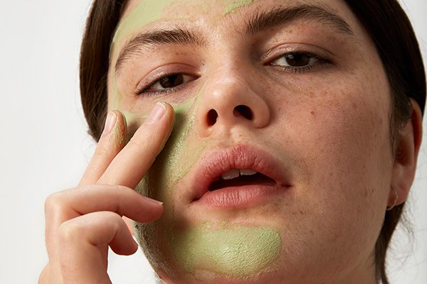 a close up image of a female applying a green clay mask to her skin in a studio setting, in front of a white background.
