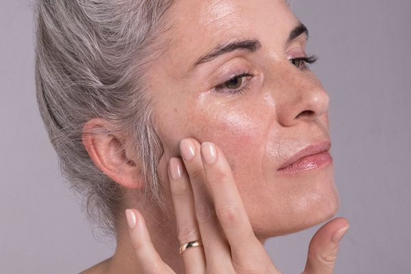 A close up image of a mature woman rubbing moisturiser into her skin, in a studio setting in front of a grey background.