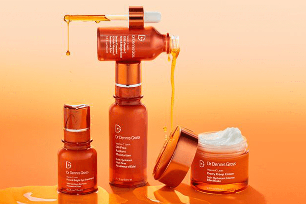 4 different Dr Dennis Gross products stacked up on one another against an orange background. two of the bottles are open and the contents leaking