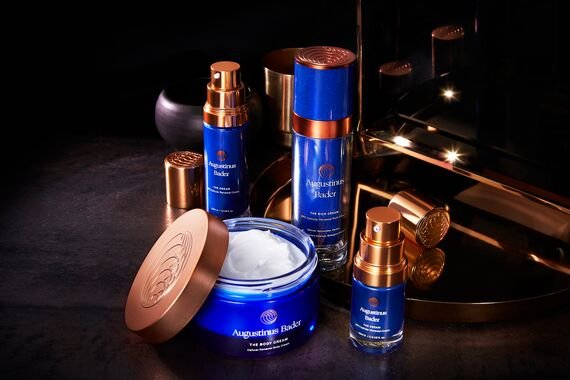 a collection of blue augustin bader skin care products against a glowy black background