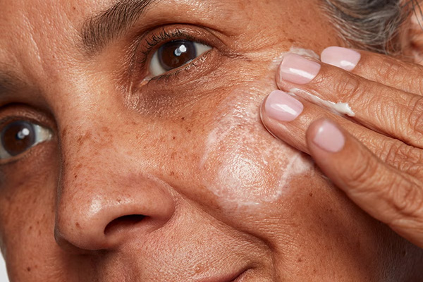 A close up image of a older female model applying a peptide serum to her skin.