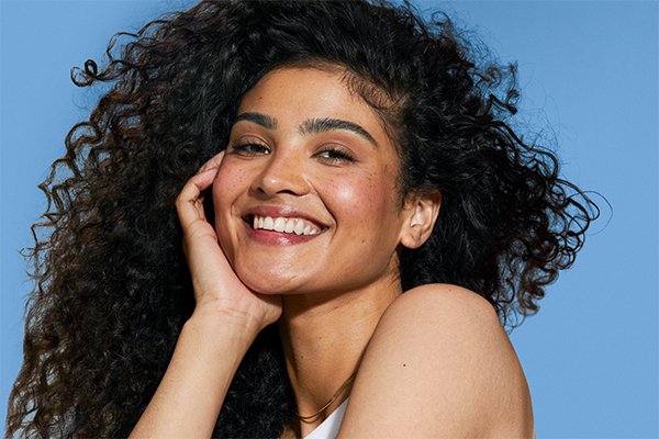 medium skinned model with big bouncy hair smiling against a blue back drop