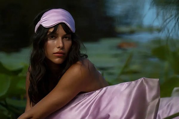 model with a pink slip silk eye mask on her head sitting near a pool of lilypads with a pink silk pillowcase on her lap