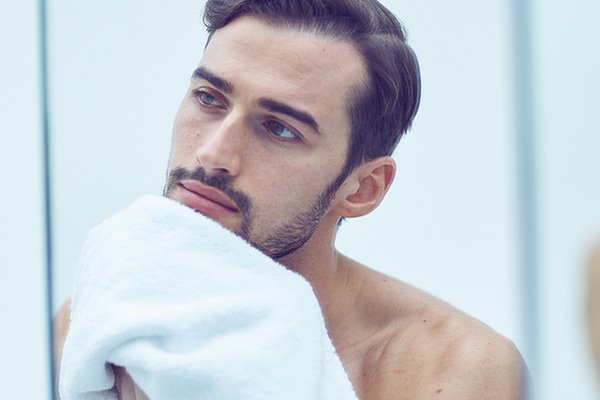 A medium shot of male model with dark hair wiping his face with a tile after his grooming routine in a bathroom setting. 