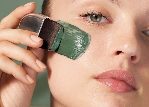 A close up of a female model applying a green clay mask to her face with brush in a studio setting in front of a green background.