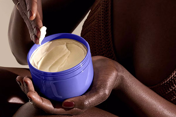 A close up of a female model with deep toned skin applying Sol de Janeiro Delicia Drench Body Butter to her legs. 