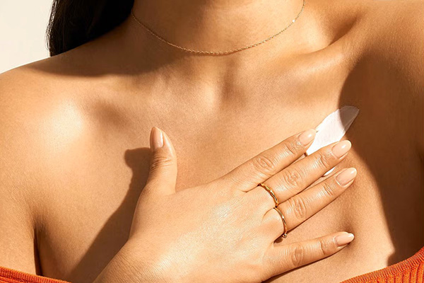 live skinned model applying something white to her chest. you cannot see her face only her chest and hands. shes wearing a thin necklace and dainty rings 