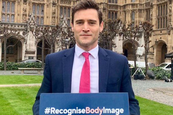 Dr Luke Evans outside parliament holding a #recognisebodyimage placard 