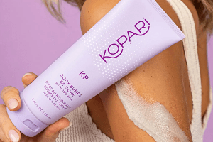model holding a kopari beauty body bumps be gone with some of the product on the back of her arm against a purple background