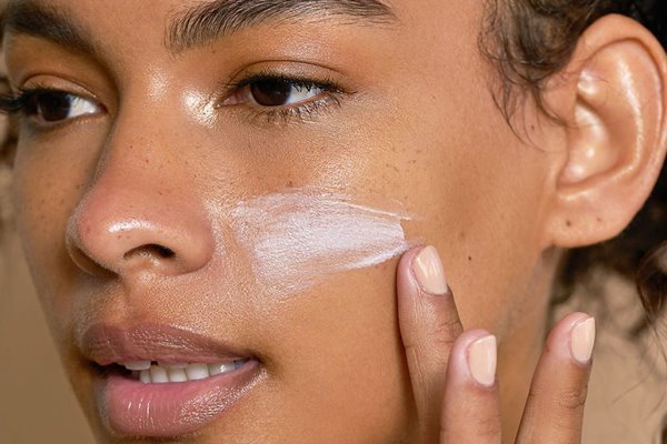 A close up of a bare-faced model with curly hair applying sunscreen on her cheek in a studio setting with a brown background