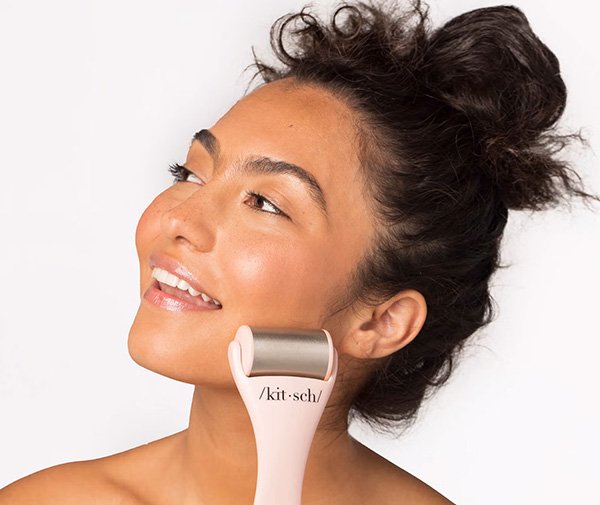 A medium shot of a female model using Kitsch's face roller over her face and smiling on a white background in a studio setting.
