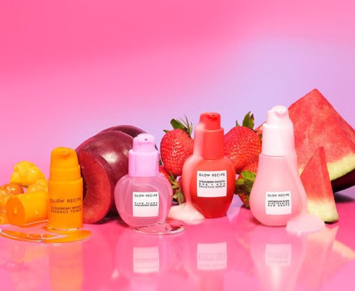 A selection of Glow Recipe products in a studio setting against a pink and purple background with fresh fruit including plums.