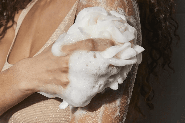 Close up of woman cleansing her body in the shower with a foam cleanser, loofa and curly hair in a bathroom setting. 