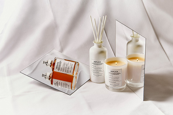 A wide shot of multiple Maison Margiela Replica fragrances – including a perfume, candle and diffuser – layer on white cloth and mirrors.