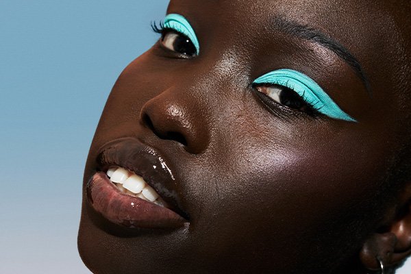 A close up of a deep-skin toned model, wearing bright blue eyeshadow in a studio setting, in front of a blue background. 