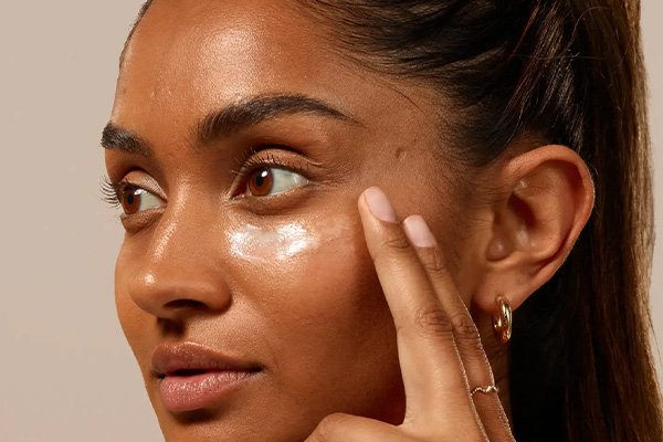 Young female model looking away from the camera applying coral reef safe eye cream to under eyes in a studio setting with brown background.