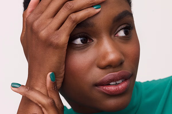 A close up of a deep-toned model wearing a green rollneck top and matching green nail varnish. In a studio setting, in front of a white background.