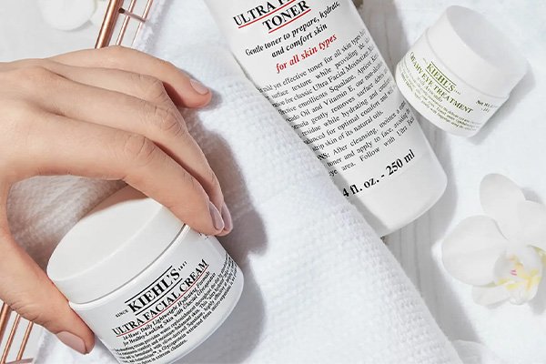 A hand picking up the Kiehl’s Ultra Facial Cream and adding it to a basket of other Kiehl’s products including a toner and eye cream.