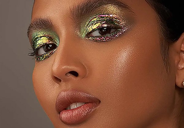 Woman of colour wears purple and green holographic eye make up against a studio background