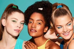 one brunette model with her hair up, one darker skinned model with curl hair tied up and one blonde model with her hair tied up smiling, shot in a studio with a green background