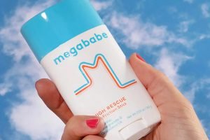 a models hand with painted red nails holding a megababe thigh rescue bottle against a blue sky with clouds