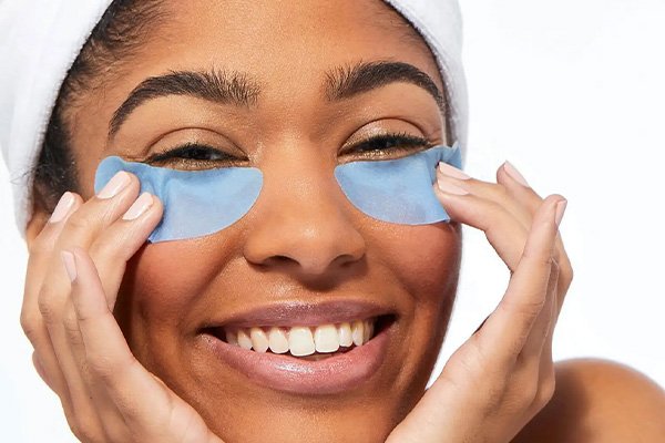 Medium shot of dark skinned model smiling at the camera wearing a head towel and blue eye mask on her undereyes, in a bathroom setting. 