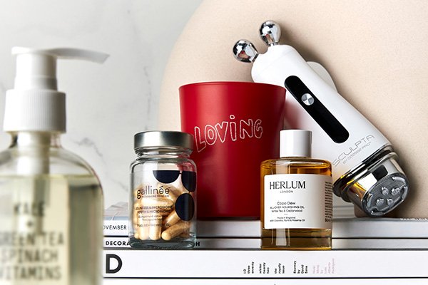 An assortment of beauty products including a skin care tool, a cleanser, a red candle, a jar of supplements, and a skin oil.