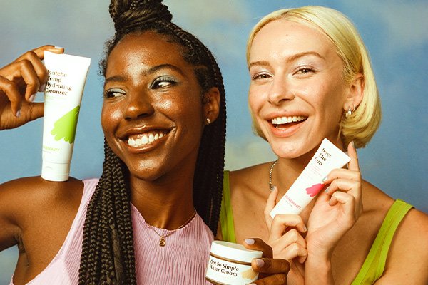 A wide shot of two models, one blonde in a green top and one black haired in a pink top, holding KraveBeauty skin care while smiling. 