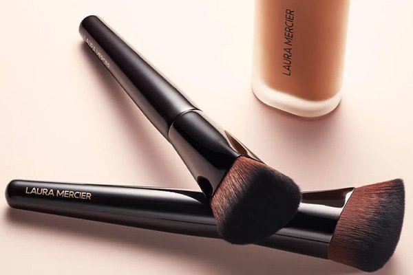 A wide shot of two Laura Mercier make up brushes stacked on top of each other with the foundation in the background, in a studio setting.