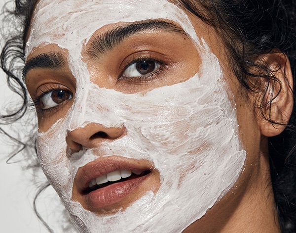 Model with tanned skin and curly hair wears a skin care mask on her face