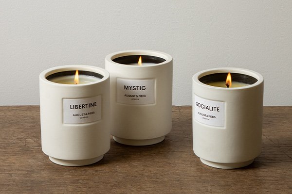 A lifestyle wide shot of three AUGUST&PIERS candles lit up on a wooden table top with a grey background in a studio setting. 