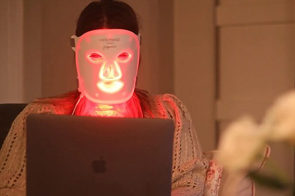 Woman wearing a Deesee Pro LED face mask that is reflecting red light on the sofa while typing on her laptop in a living room setting. 