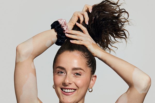 A medium shot of a brunette model with curly, long, black hair tying up her hair while smiling in a studio setting, on a grey background.