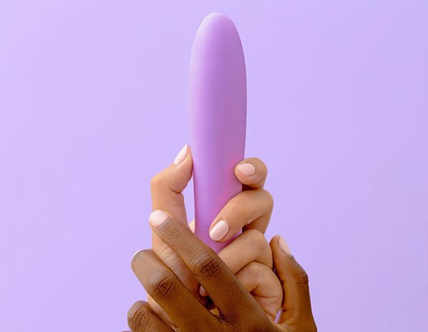 A wide shot image of two hands of different skin tones holding a purple Smile Maker’s vibrator, in a studio setting on a purple background.